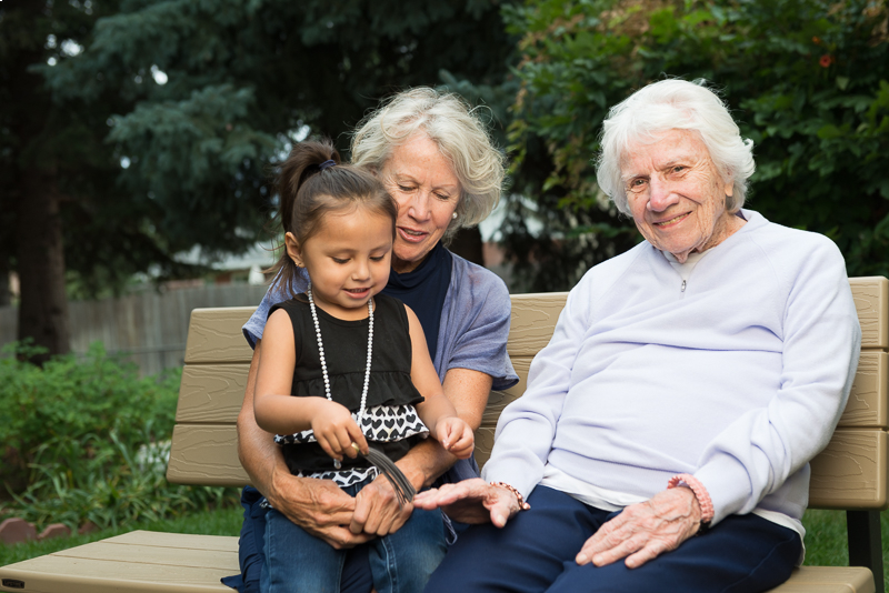 Three generations of women sitting on a park bench; a girl is showing a chain to her great-grandmother.