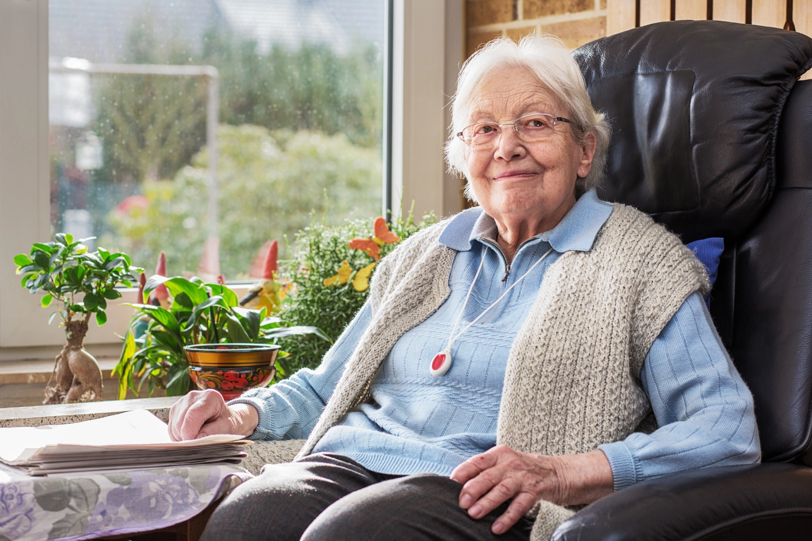 Elderly woman wearing a medical alert necklace and smiling while sitting in a black armchair.